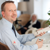 Foto: Ein Mann in DRK-Hemd wechselt die Batterie einer Fernbedienung aus - im Hintergrund eine Seniorin.